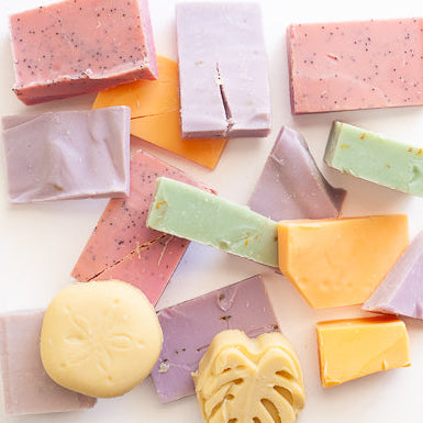 A selection of soap bars of different colors (pink, orange, light green, light purple, yellow) and shapes (split rectangles, slightly smashed monstera leaf, sand dollar, triangles) that are all slightly damaged with cracks, dents or other inconsistencies.