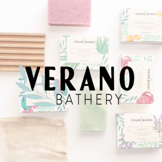 A flat-lay arrangement of Verano Bathery products on a white background, including several botanical-patterned soap bars, a wooden soap dish, a beige washcloth, and a central black text logo reading 'VERANO BATHERY'.